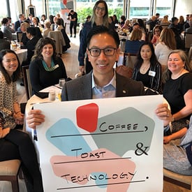 A man standing in front of a room full of event attendee holding a colorful sign that says: 